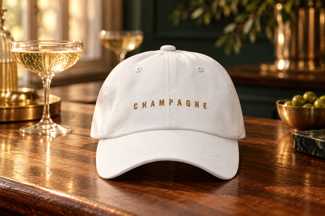 White Champagne cap with gold embroidery on a wooden cocktail bar beside sparkling wine glasses.